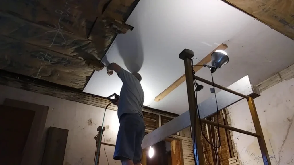 Drywall contractor installing ceiling drywall panels during a residential renovation project using professional lifting and fastening equipment.
