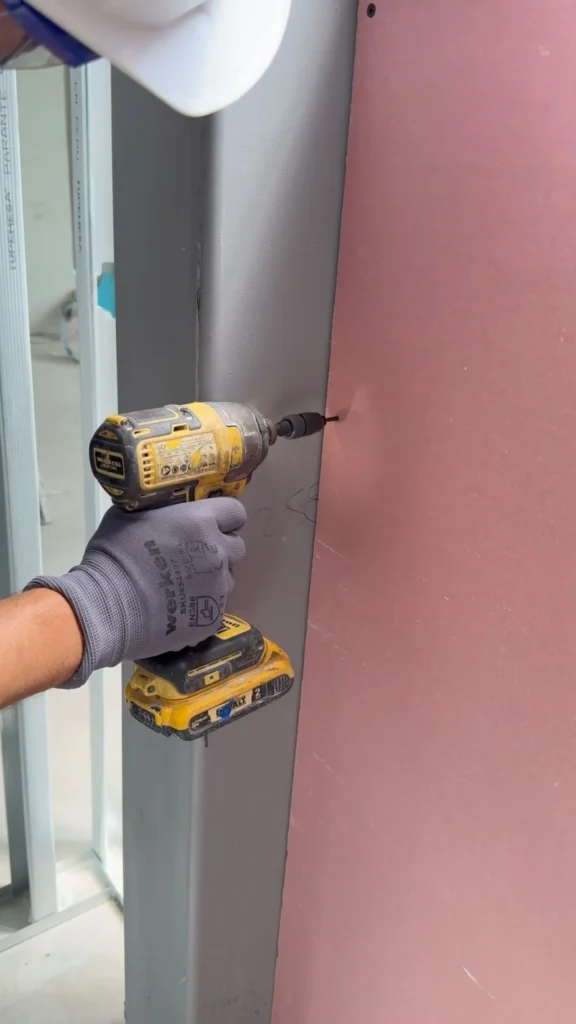 Drywall contractor fastening gypsum board to metal stud framing using a power drill during commercial wall installation.