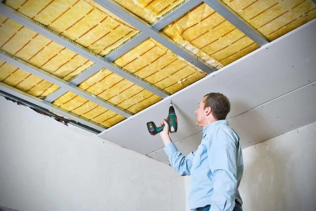 Worker installing drywall ceiling panels beneath fiberglass insulation using a power drill.