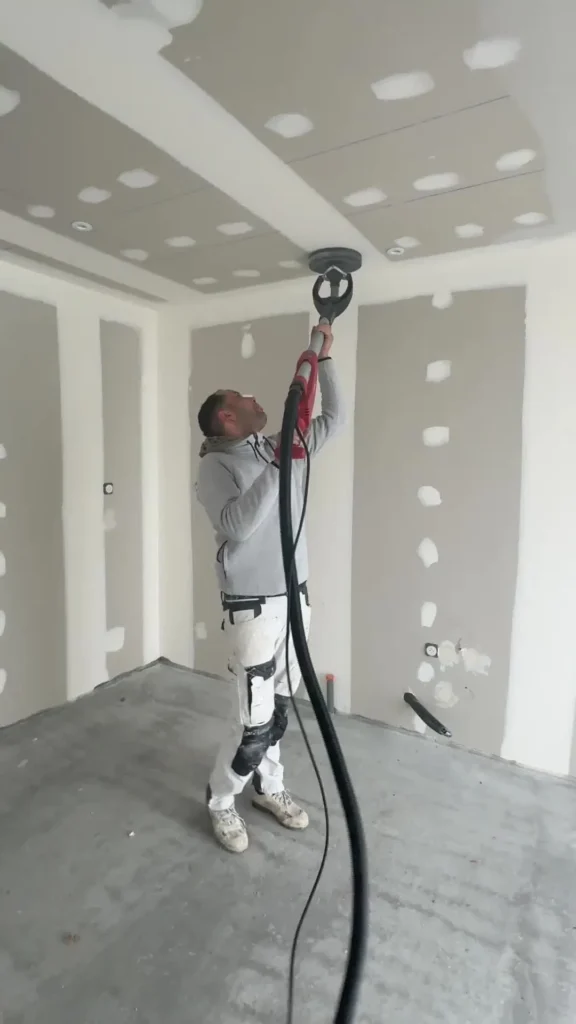 Worker sanding drywall ceiling joints with a power sander during interior finishing.