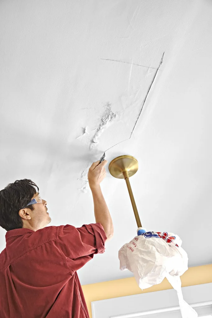 Water-damaged ceiling drywall being cut and repaired around a crack near a ceiling light fixture.