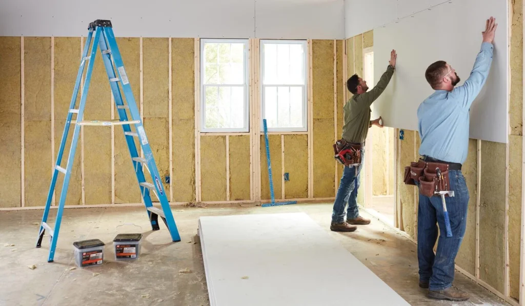 Professional drywall installers mounting wall panels in a residential construction project with insulated framing and interior finishing work.